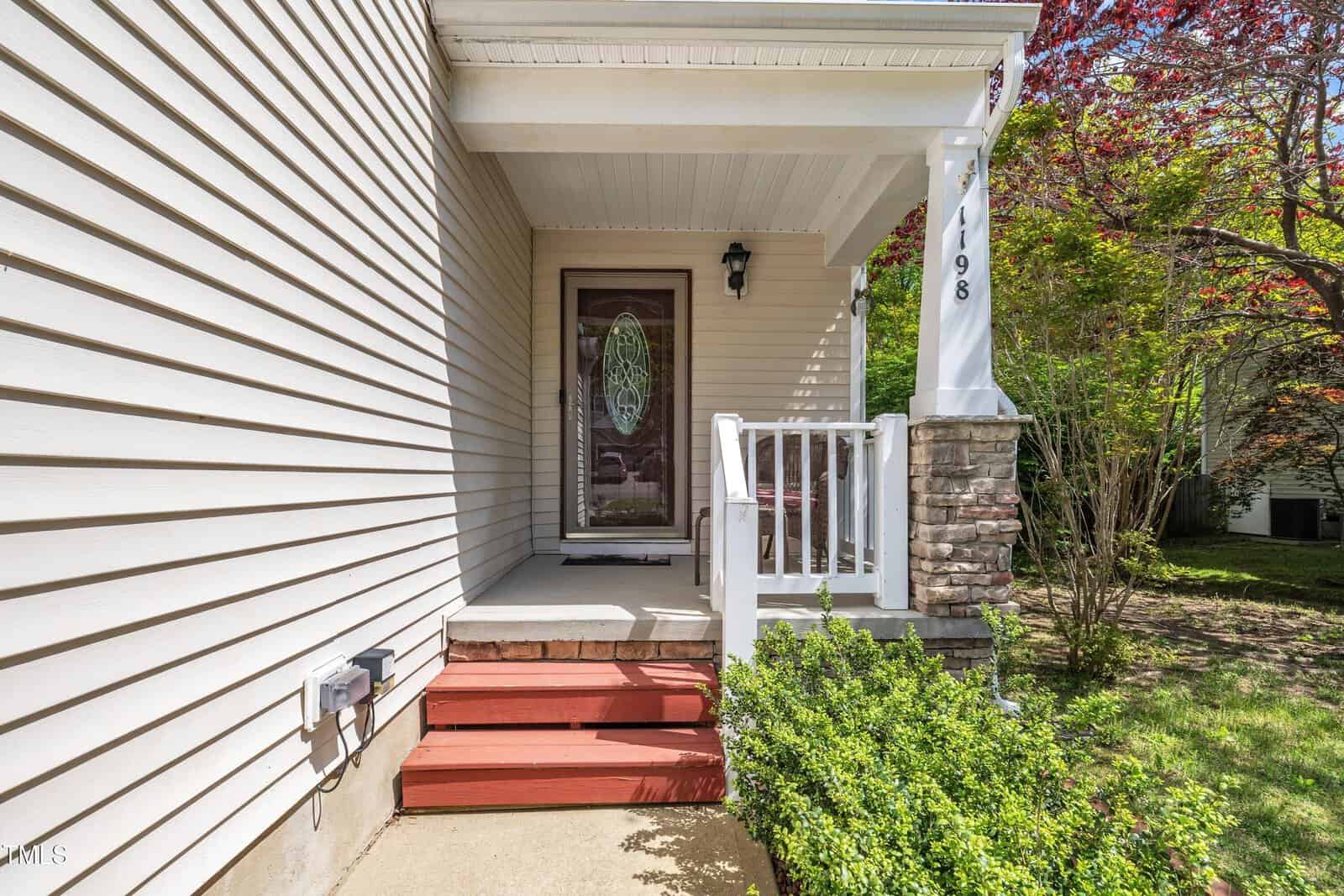 The image shows a small covered front porch with red steps, a white railing, and a glass front door. The house exterior is beige siding with a stone column, and there are shrubs and grass next to the walkway.