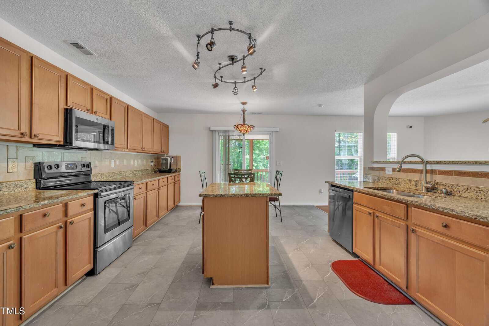 Spacious kitchen with wooden cabinets, stainless steel appliances, center island, granite countertops, tile floor, and a modern light fixture; dining area visible near glass doors leading outside.