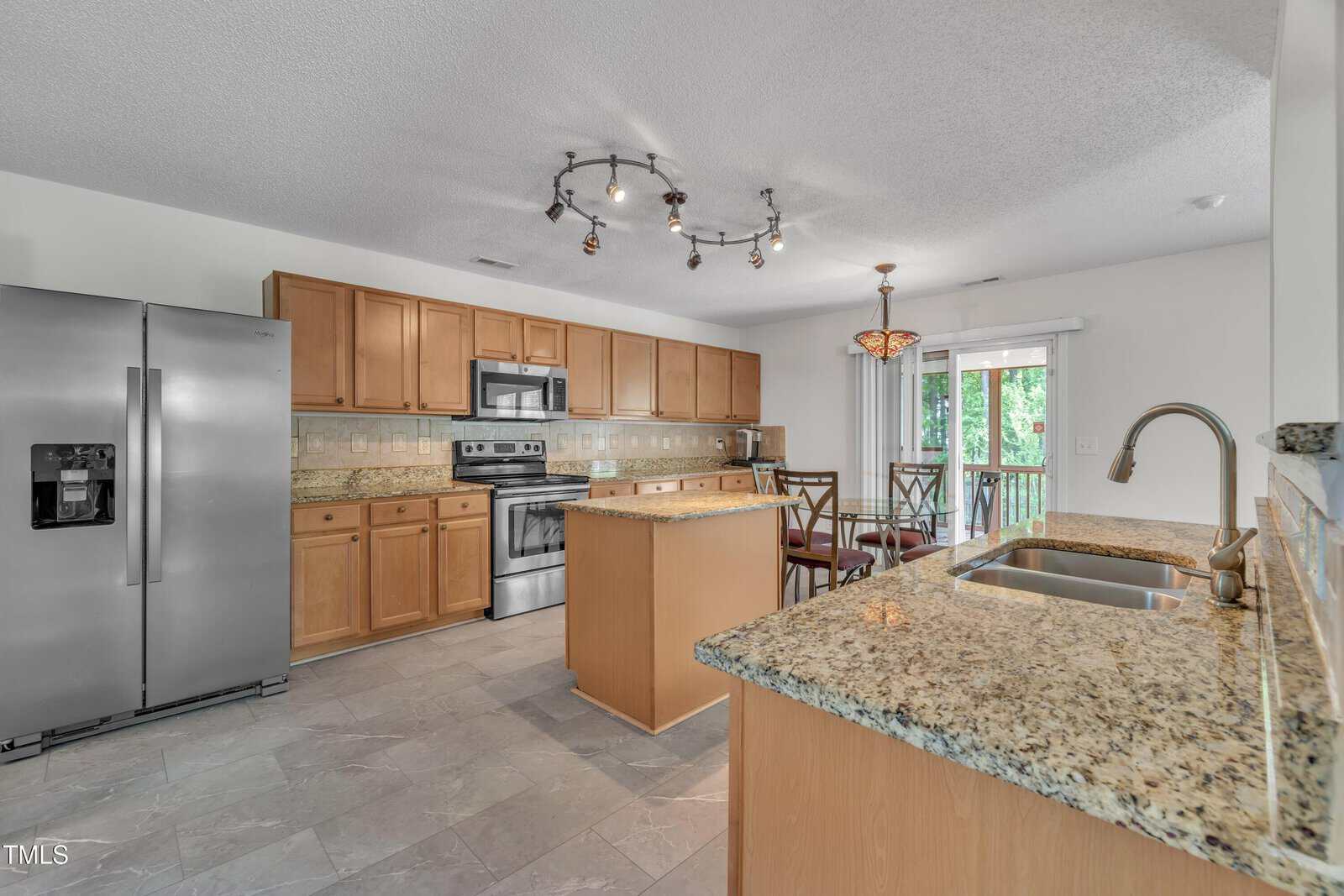 A modern kitchen with stainless steel appliances, wood cabinets, granite countertops, a central island, and a dining area beside sliding glass doors leading to a balcony.