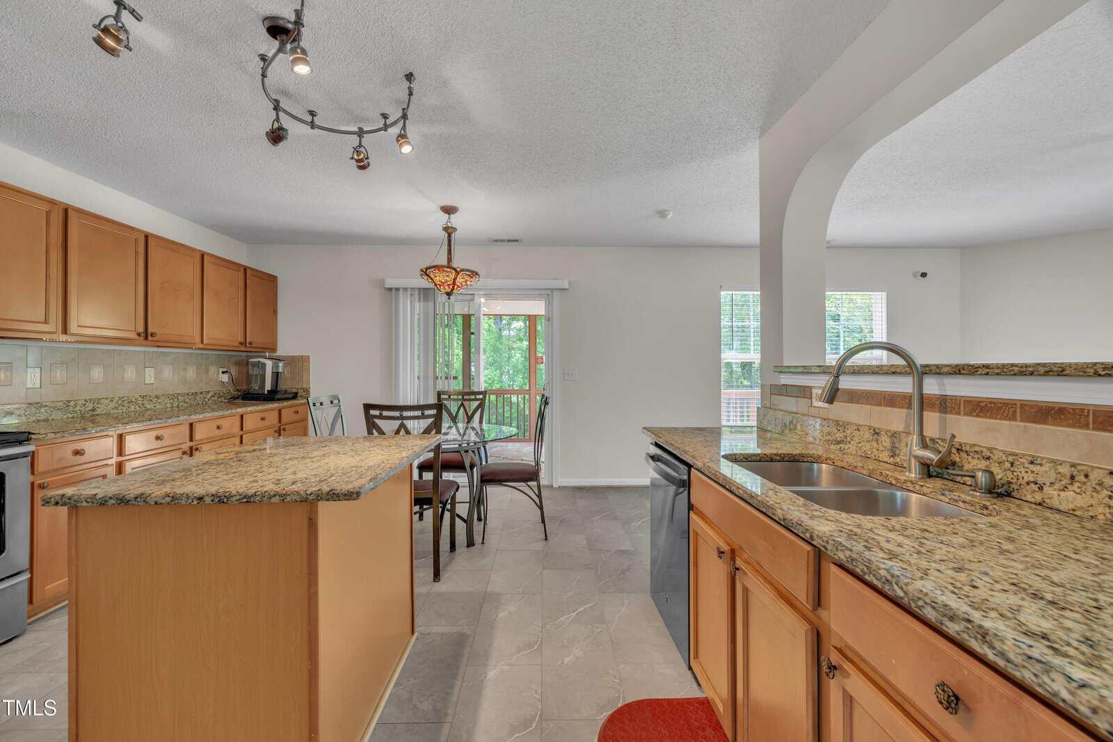 Modern kitchen with light wood cabinets, granite countertops, a center island, stainless steel appliances, tile flooring, a double sink, and a dining area with sliding glass doors leading to a balcony with greenery visible outside.