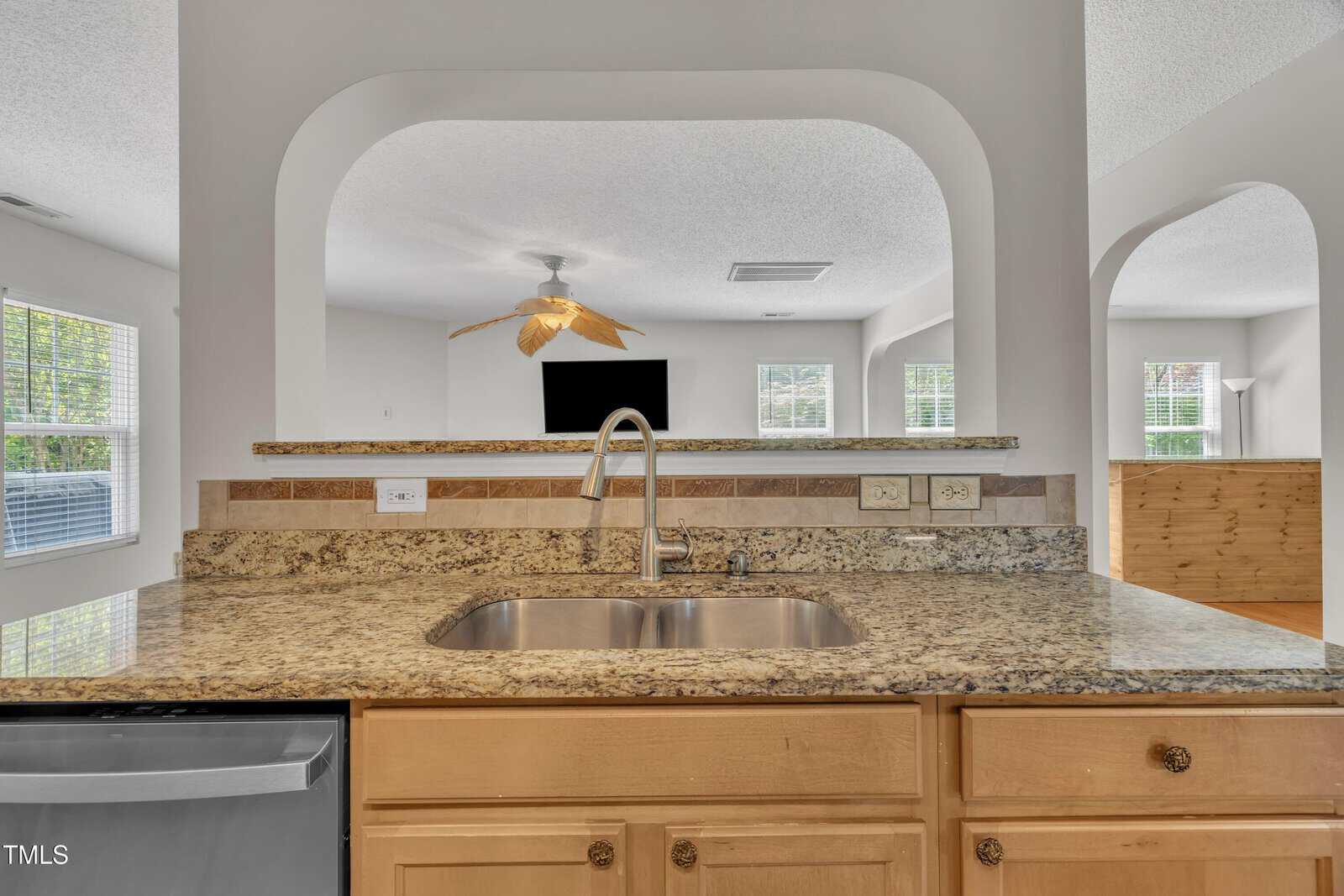 A granite kitchen countertop with a stainless steel sink and faucet, light wood cabinets, and a dishwasher. In the background, a living area with large windows, a ceiling fan, and a wall-mounted TV is visible.
