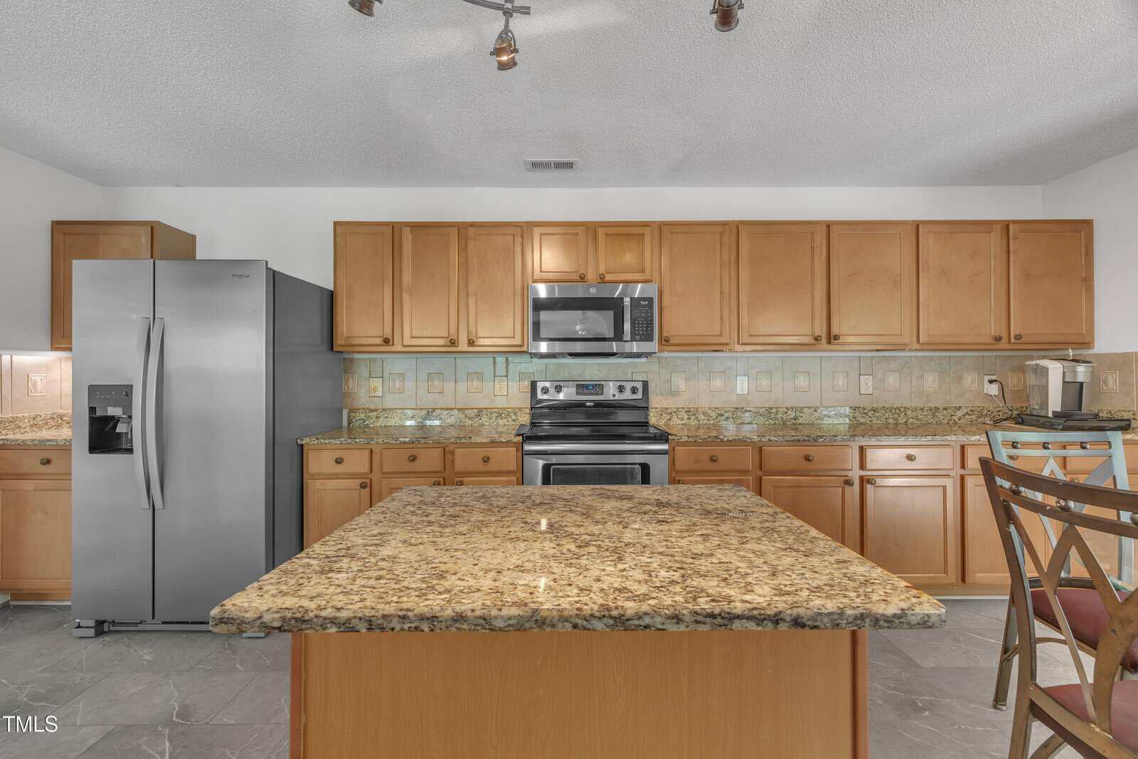 A modern kitchen with wooden cabinets, stainless steel appliances including a refrigerator, oven, and microwave, and a large granite island in the center. There are two chairs at the island and a coffee maker on the counter.