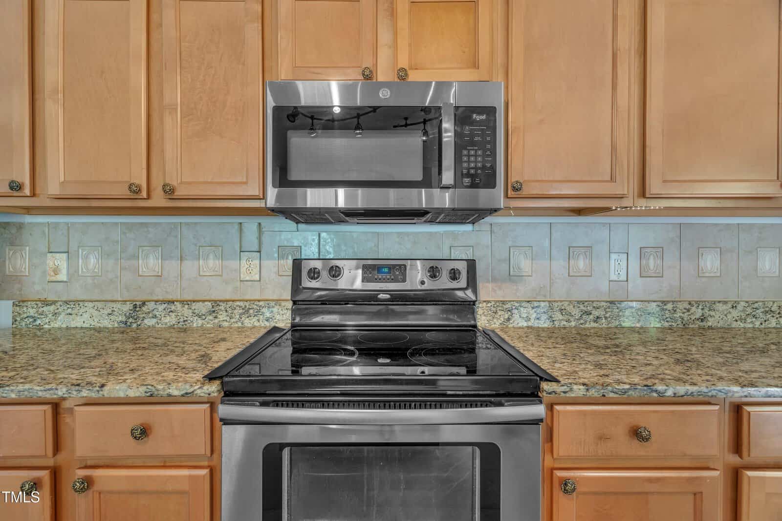 Stainless steel microwave and electric stove with black cooktop set against a granite countertop and light wood cabinets; beige tile backsplash with decorative accents visible in the background.