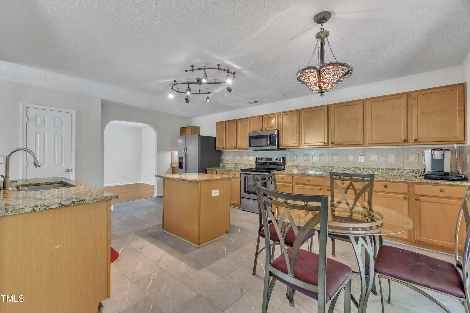 A modern kitchen with light wood cabinets, stainless steel appliances, granite countertops, a small island, a round glass dining table with four chairs, and tile flooring under track and pendant lighting.