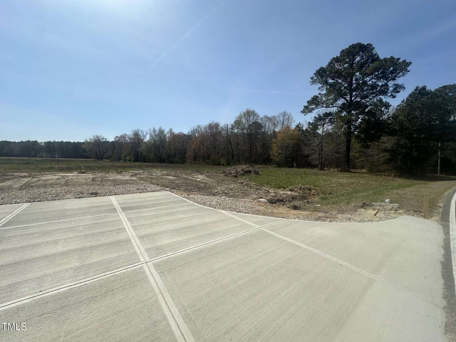 A newly paved concrete road leads to an open, grassy plot bordered by trees under a clear blue sky. Dirt and scattered brush separate the road from the wooded area in the background.