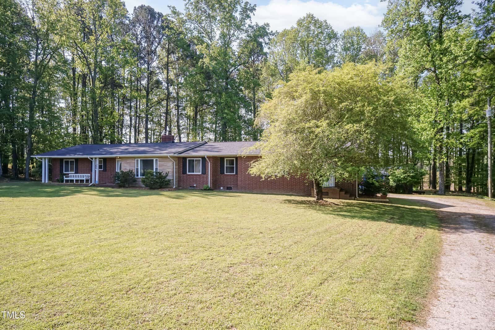 Single-story brick house with a large, neatly mowed front lawn, surrounded by tall trees. A gravel driveway leads to a carport on the right side of the house. The setting appears peaceful and wooded.