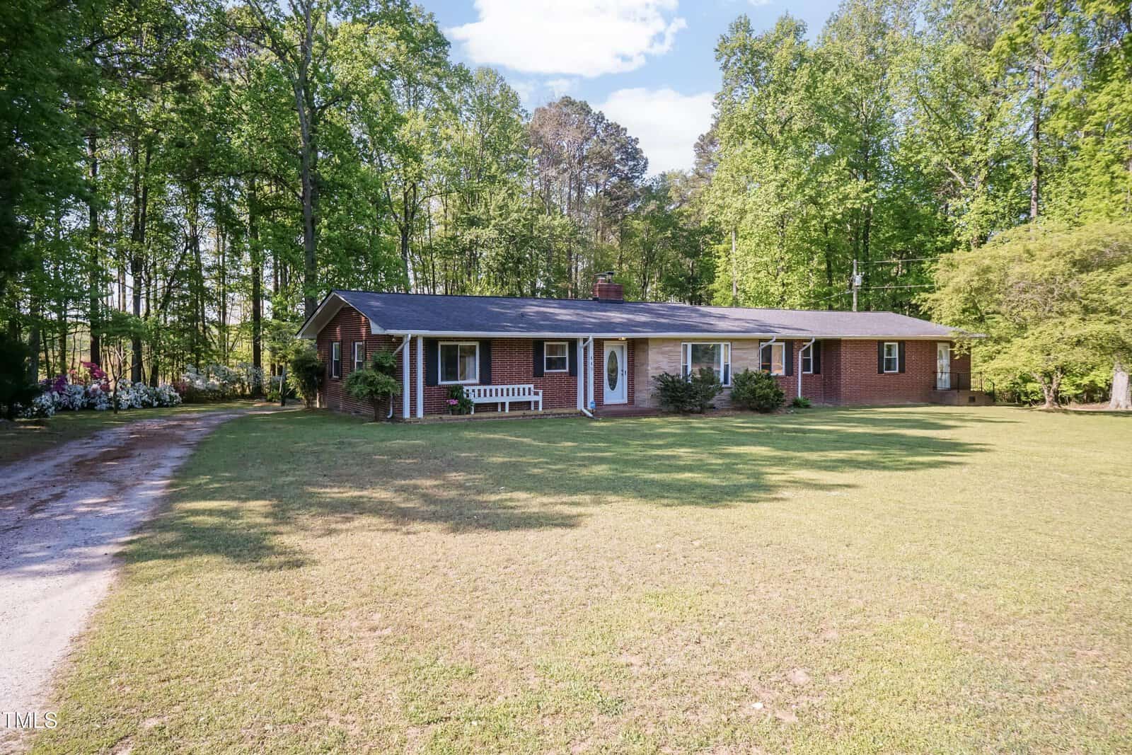 Single-story red brick house with a dark roof, white trim, and chimney, surrounded by green trees and a large grassy yard. A gravel driveway curves along the left side of the property.