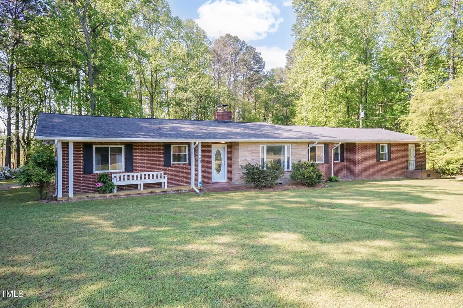 Single-story brick house with a dark roof, large front lawn, a white bench near the door, and surrounded by tall green trees under a partly cloudy sky.