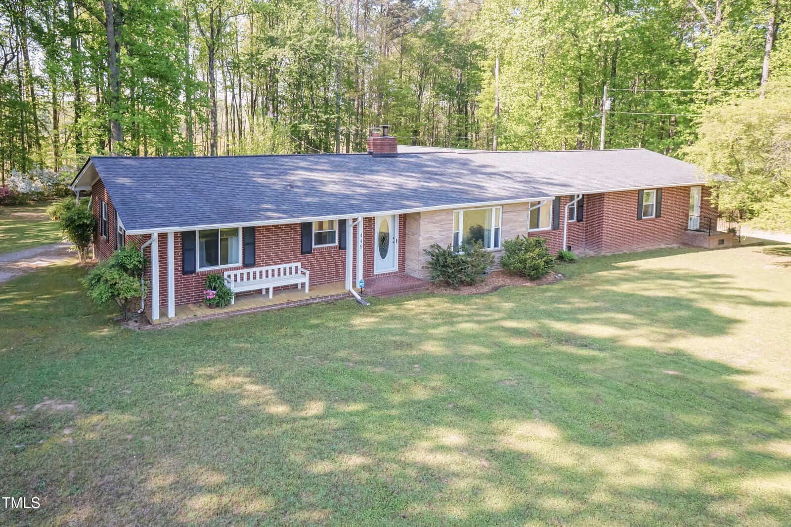 A single-story brick house with a dark shingle roof, front porch bench, several windows, and shrubs. The house is surrounded by grass and trees, with a wooded area in the background.