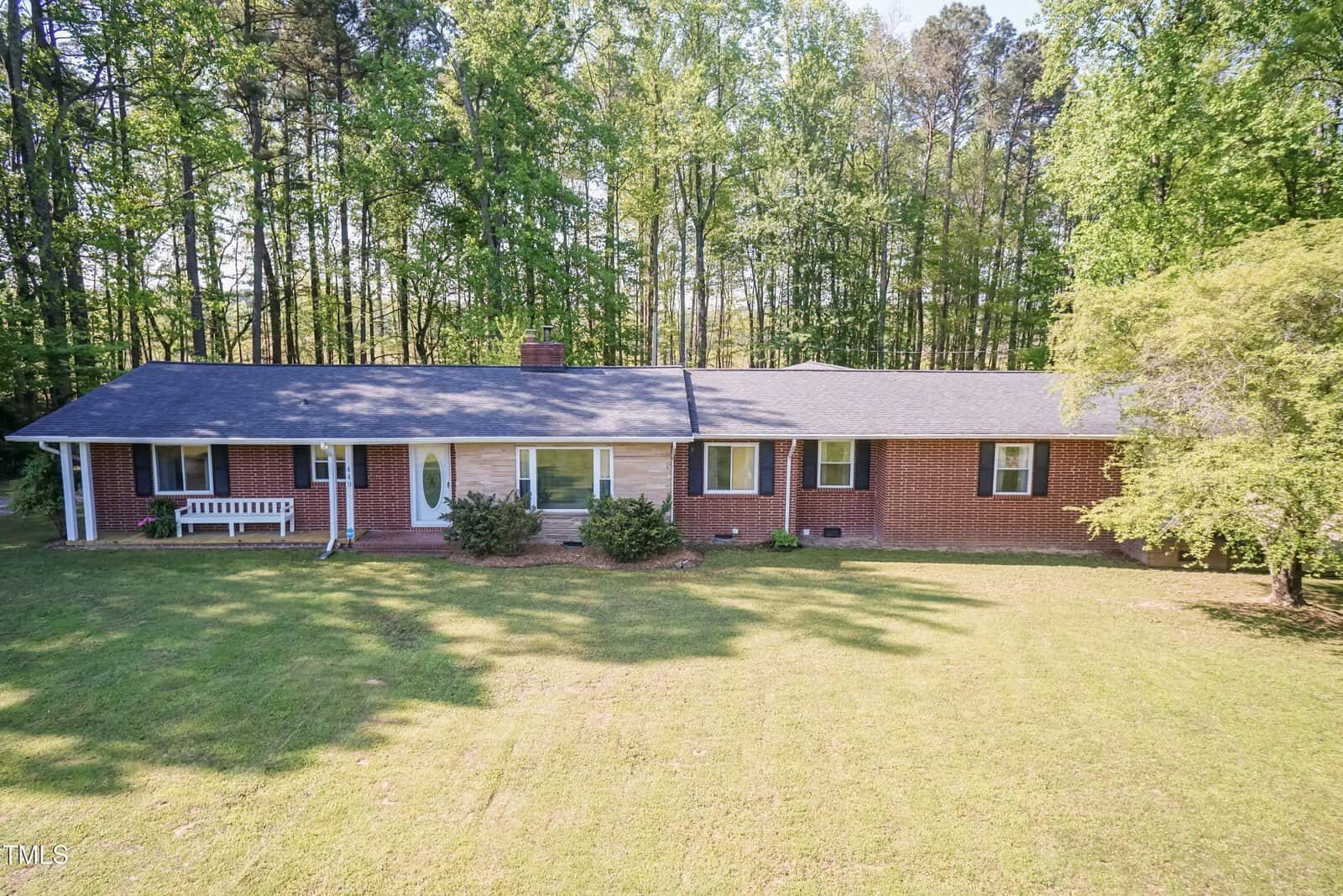 Single-story red brick house with black roof, large front lawn, white bench by the entrance, and surrounded by tall trees in the background under clear daylight.