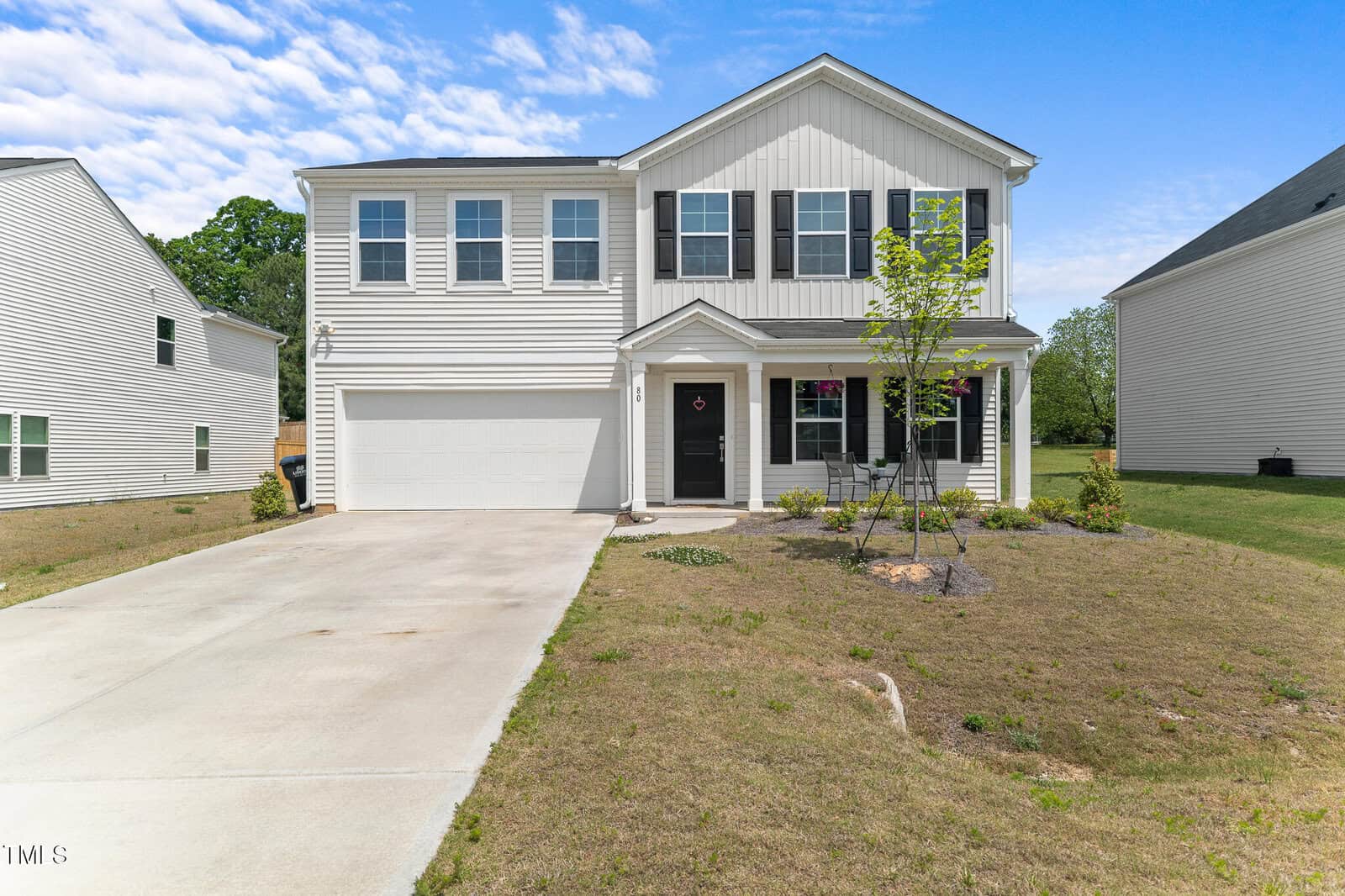 A two-story modern suburban house with white siding, black shutters, and a double garage. It has a wide driveway, small front yard with young trees, and a blue sky with scattered clouds above.