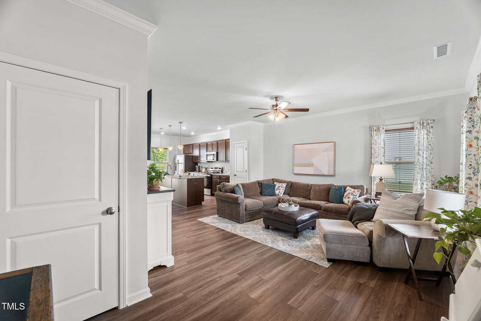 Open-concept living room with sectional sofa, patterned pillows, ottoman, and side tables, leading to a modern kitchen. Wood floors, ceiling fan, plants, and large windows with floral curtains create a cozy, inviting space.