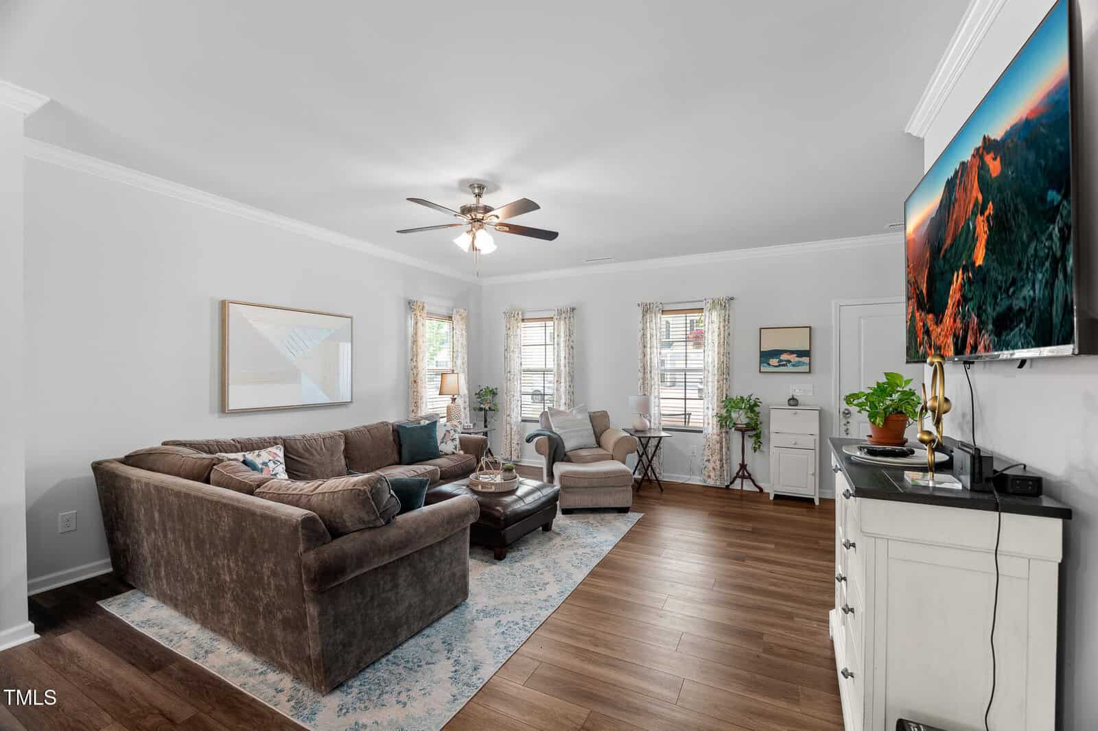 A bright living room with a brown sectional sofa, armchair, ottoman, patterned rug, wall art, potted plants, a ceiling fan, and a TV mounted on a white dresser. Three windows let in natural light.