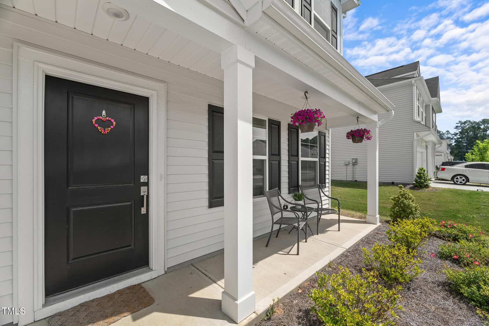A white house with a black front door adorned with a heart-shaped wreath, covered porch with two chairs and a table, hanging flower baskets, and a landscaped front yard.