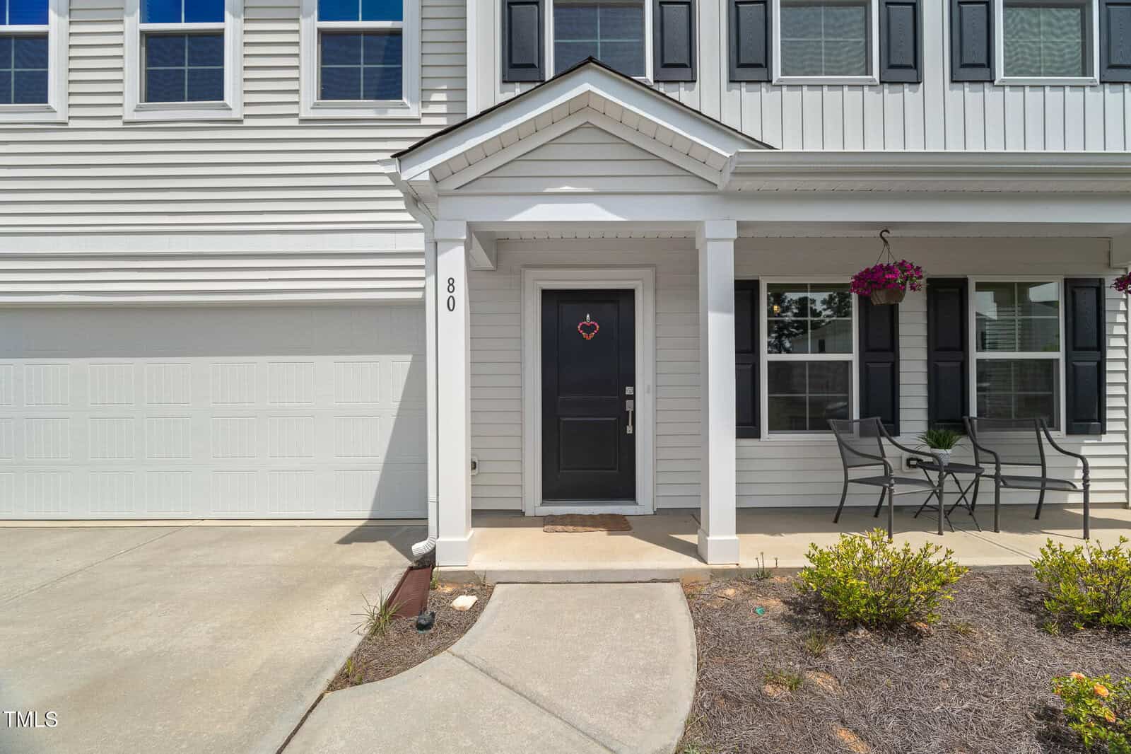 A white house with black shutters, a black door with a heart-shaped wreath, a small covered porch, two chairs with a table, hanging flower basket, and a two-car garage. Shrubs line the front walkway.