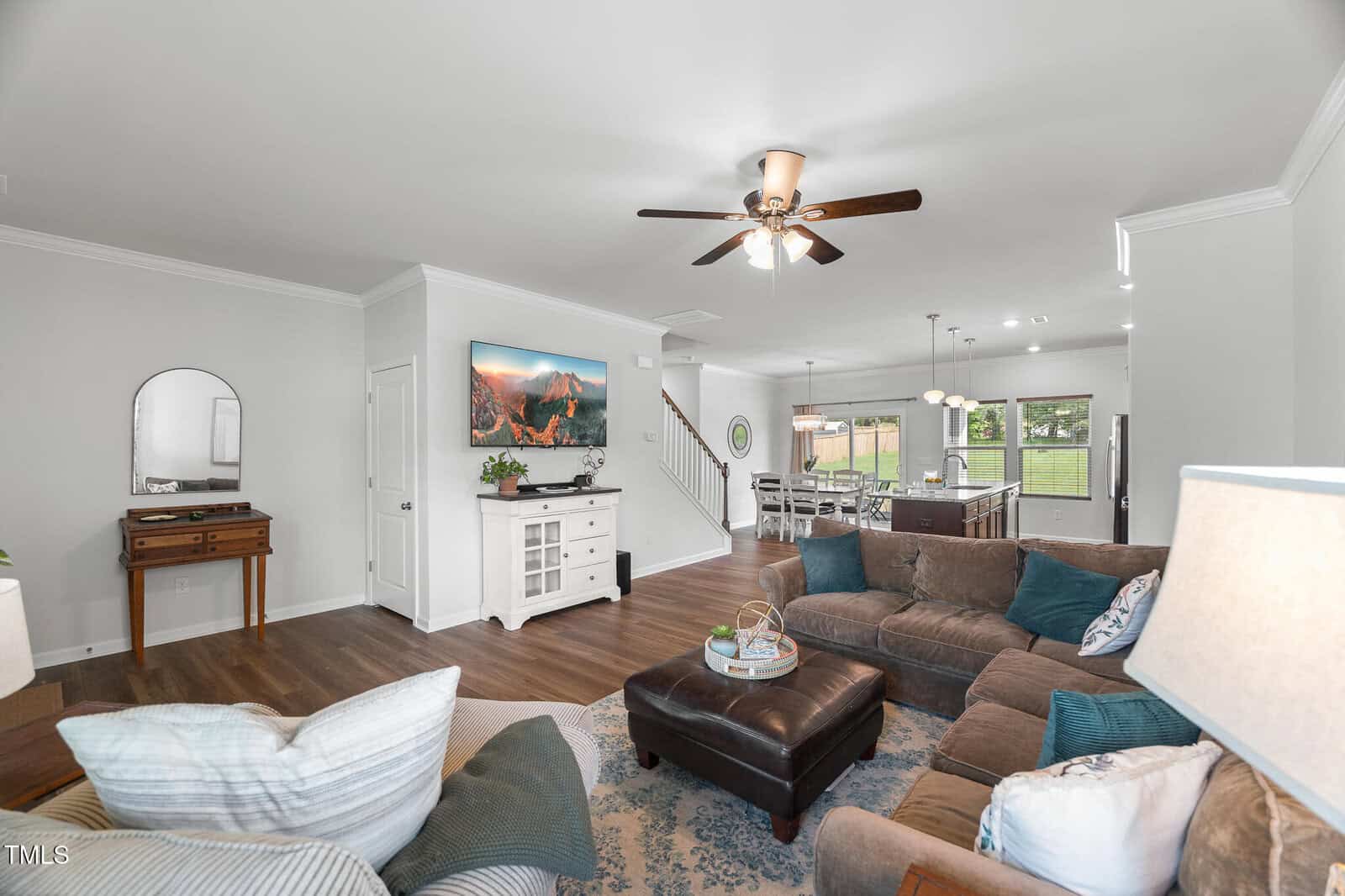 A bright living room with a brown sectional sofa, ottoman, and area rug, facing a TV on the wall. The room connects to a dining area and kitchen, with visible stairway and backyard through glass doors.