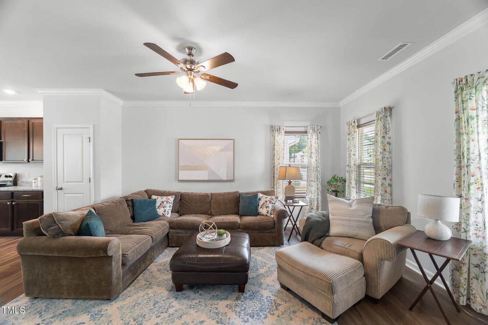 A living room with a brown sectional sofa, a matching chaise lounge, a dark ottoman, and a patterned rug. There are two windows with floral curtains, a ceiling fan, a lamp, and a framed picture on a light gray wall.