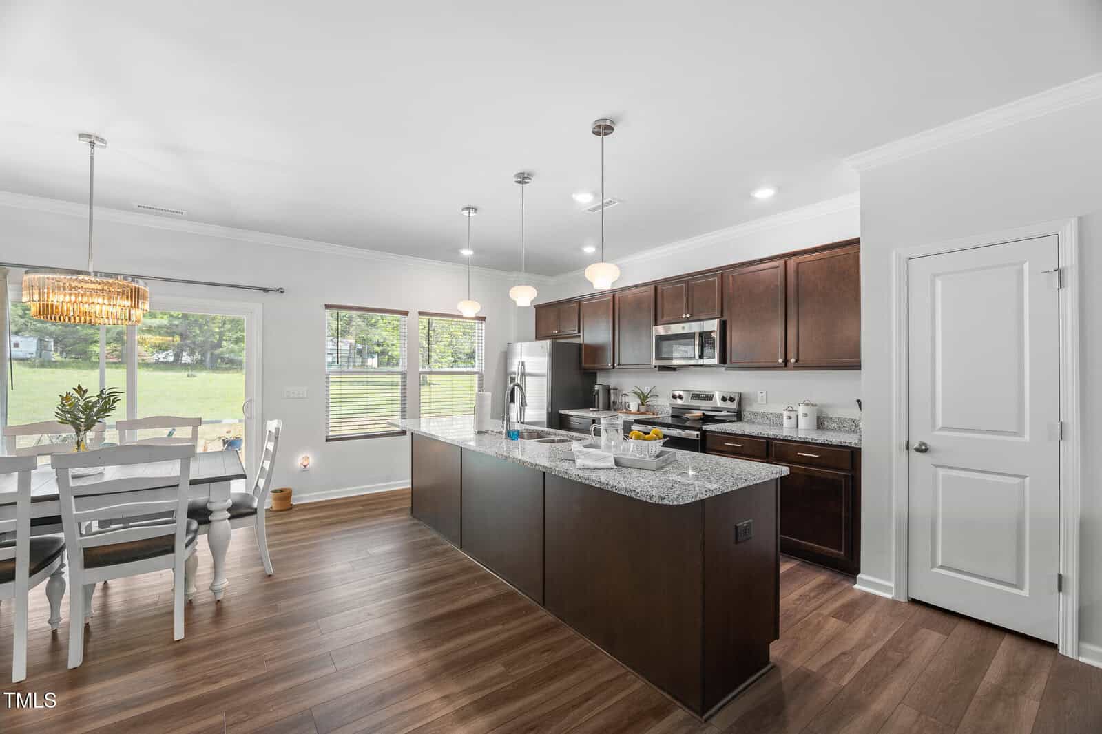Modern kitchen with dark wood cabinets, granite countertops, stainless steel appliances, and an island with sink. Dining area with white table, chairs, and large windows overlooking a green yard. Wood floors throughout.