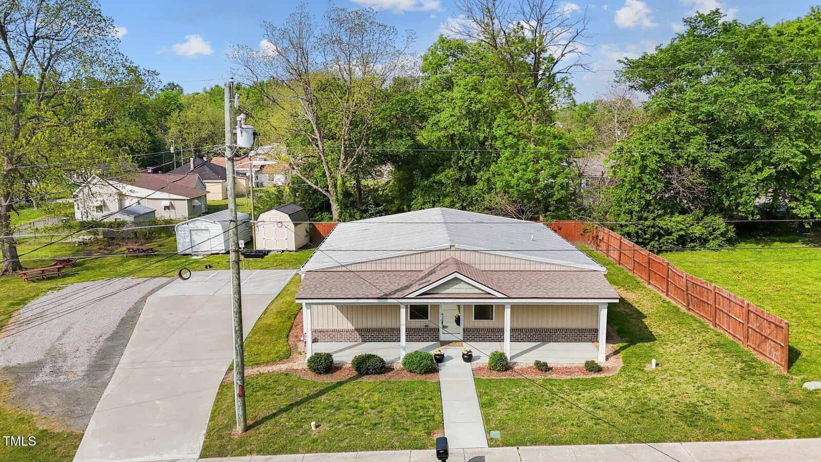 A single-story house with a covered front porch, brown shingle roof, and beige siding, sits on a grassy lot with a paved driveway, fenced backyard, and surrounding trees under a blue sky with clouds.