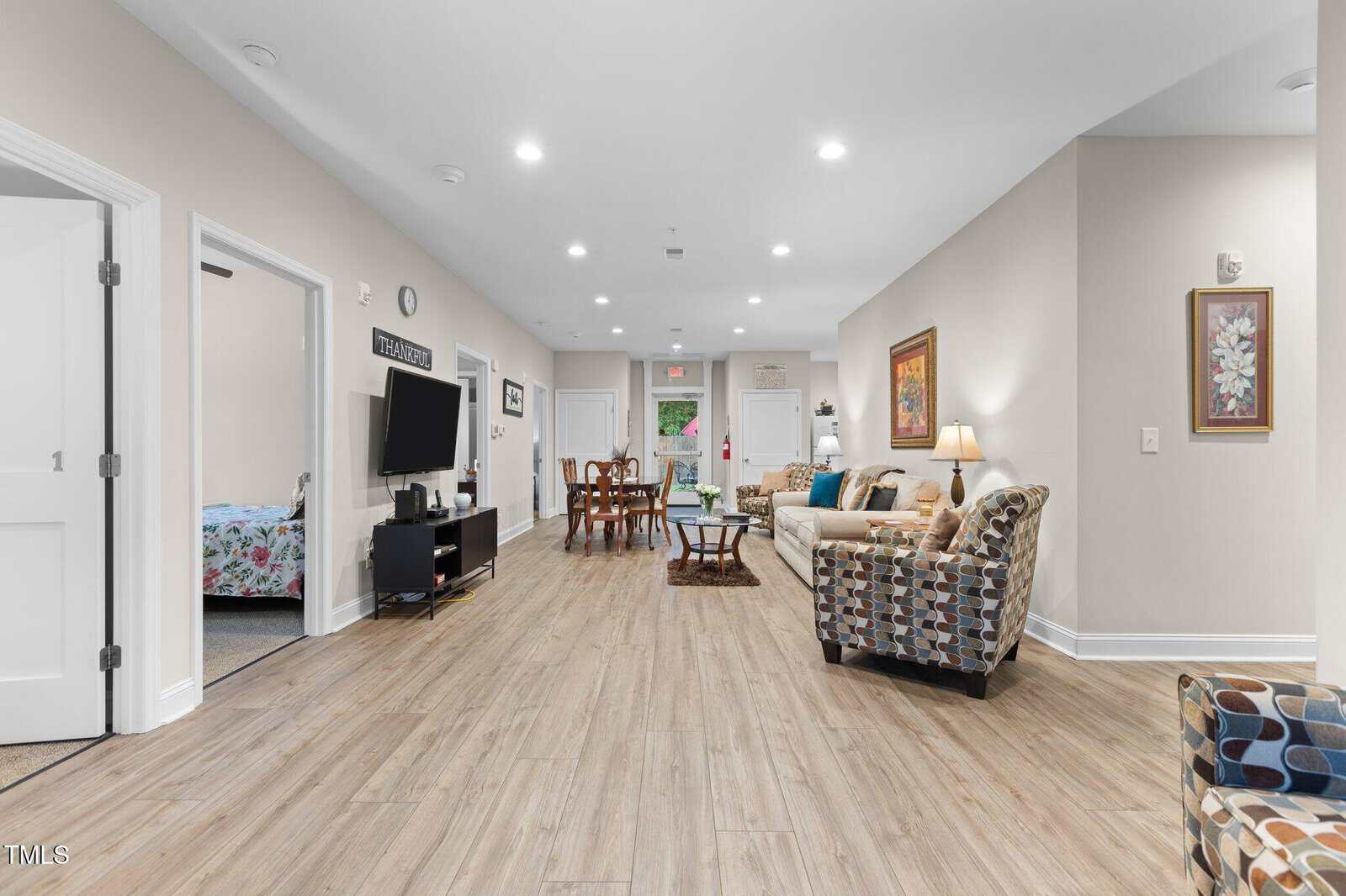 A spacious living area with wood flooring, beige walls, and recessed lighting. The room includes a seating area with couches and chairs, a TV, a dining table, and an open doorway leading to a bedroom.