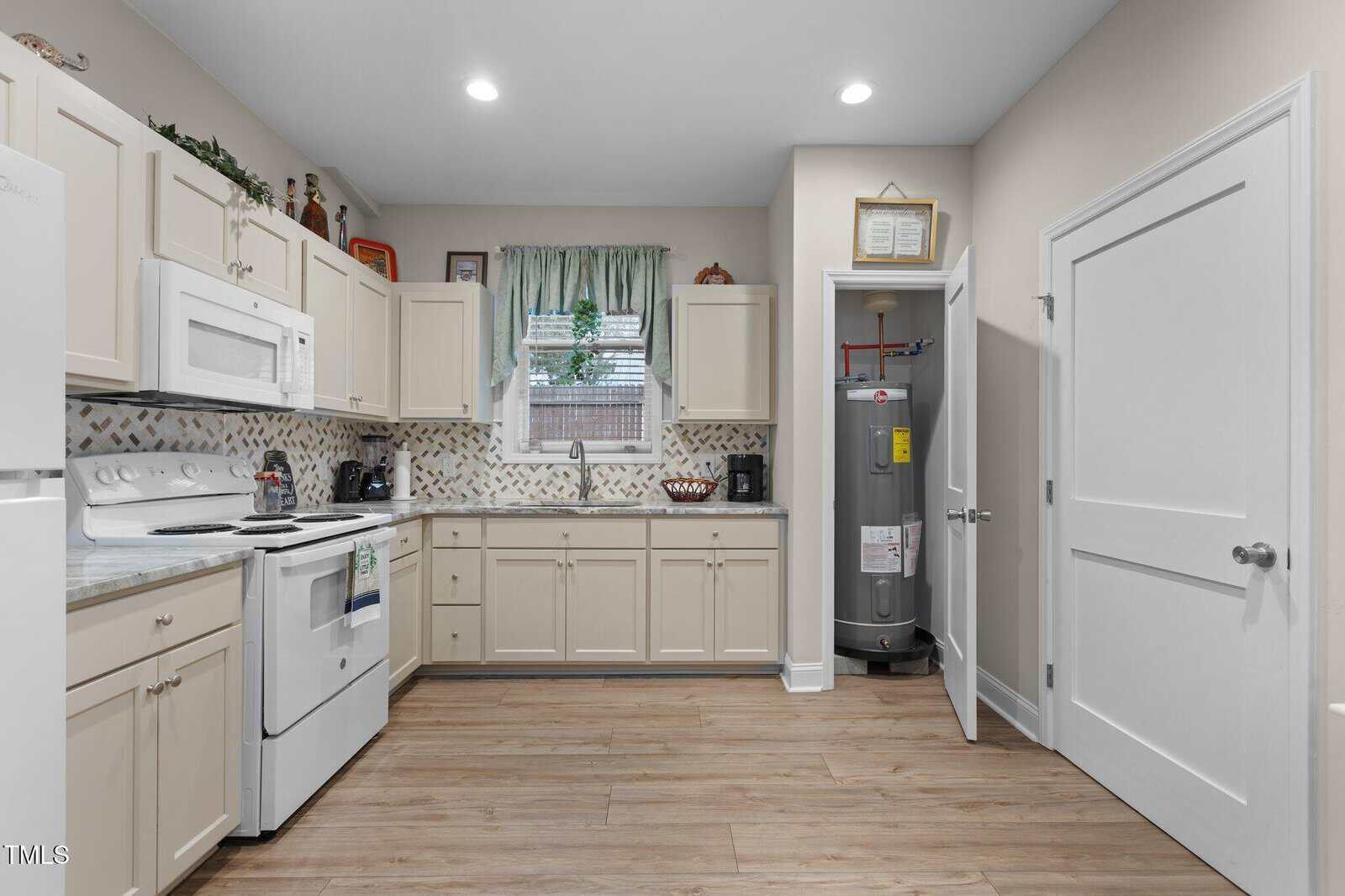 A kitchen with light-colored cabinets, white appliances, a tile backsplash, and wood flooring. A window with a green curtain is above the sink, and a water heater is visible in a corner closet with the door open.