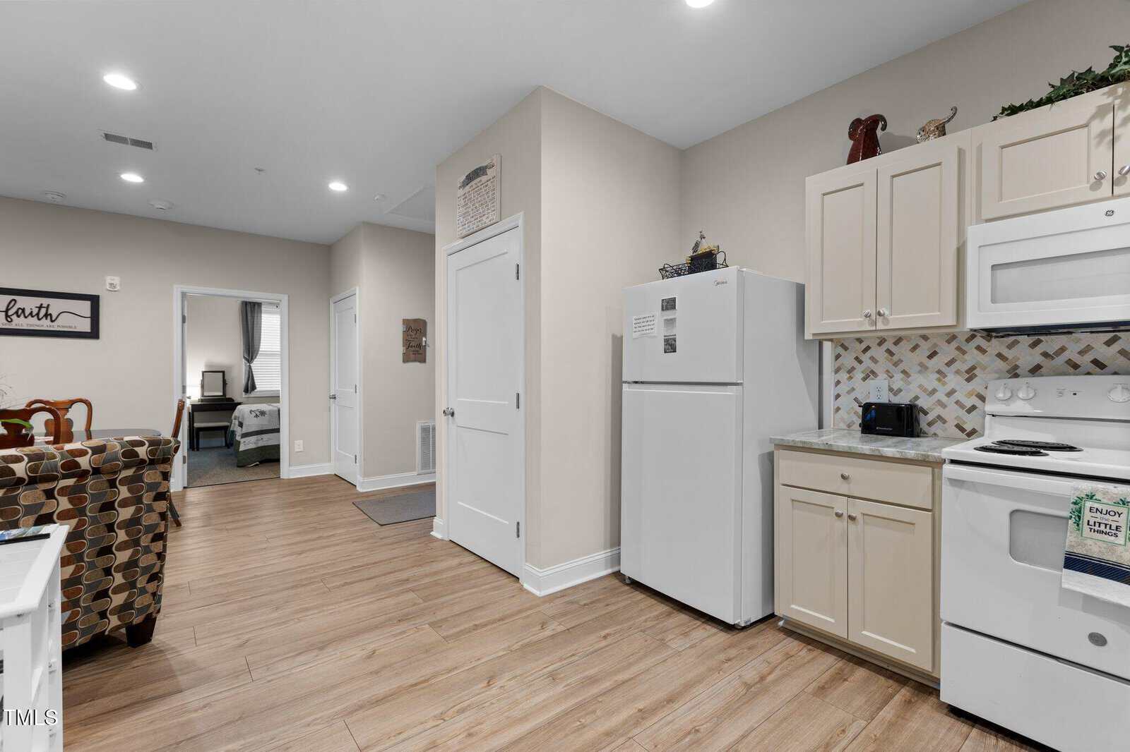 A small kitchen with white cabinets, a white refrigerator, and a white stove. The backsplash has a beige and brown pattern. Wood-look flooring extends into a living area and an open doorway leads to a bedroom.