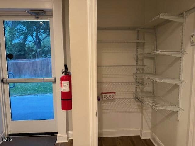 A pantry with white wire shelving is shown next to a glass exit door. A red fire extinguisher is mounted on the wall near the door. The area outside the door includes a lawn, a fence, and trees.