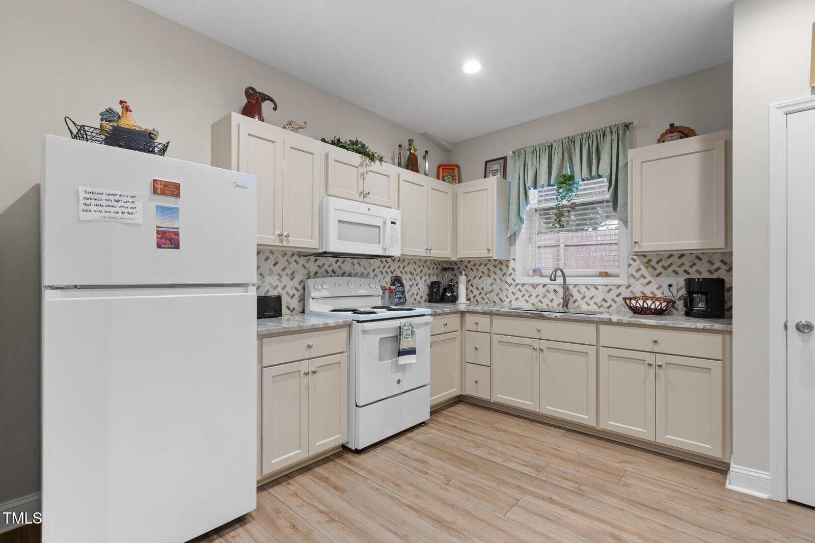 A kitchen with cream-colored cabinets, white appliances including a refrigerator, stove, and microwave, light tile backsplash, wood-look flooring, and a window with green curtains above the sink.