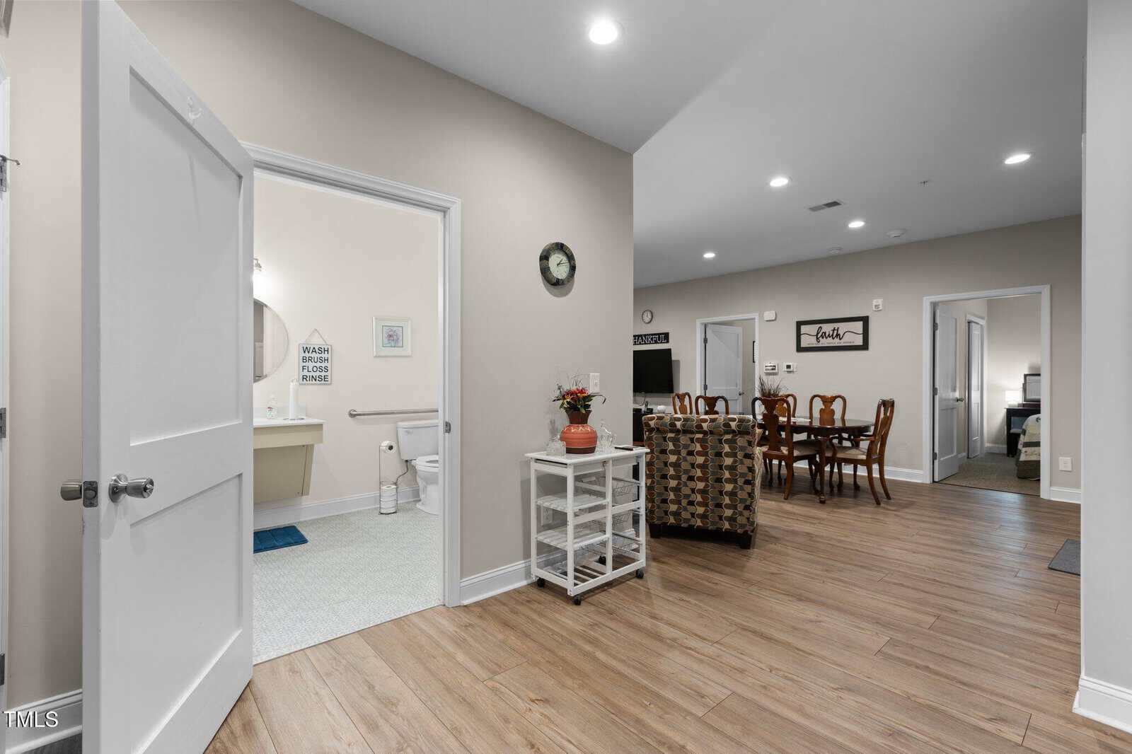 A view of a modern, accessible bathroom with grab bars and a spacious living and dining area with wood flooring, neutral walls, contemporary furniture, and decorative accents, seen from a hallway.