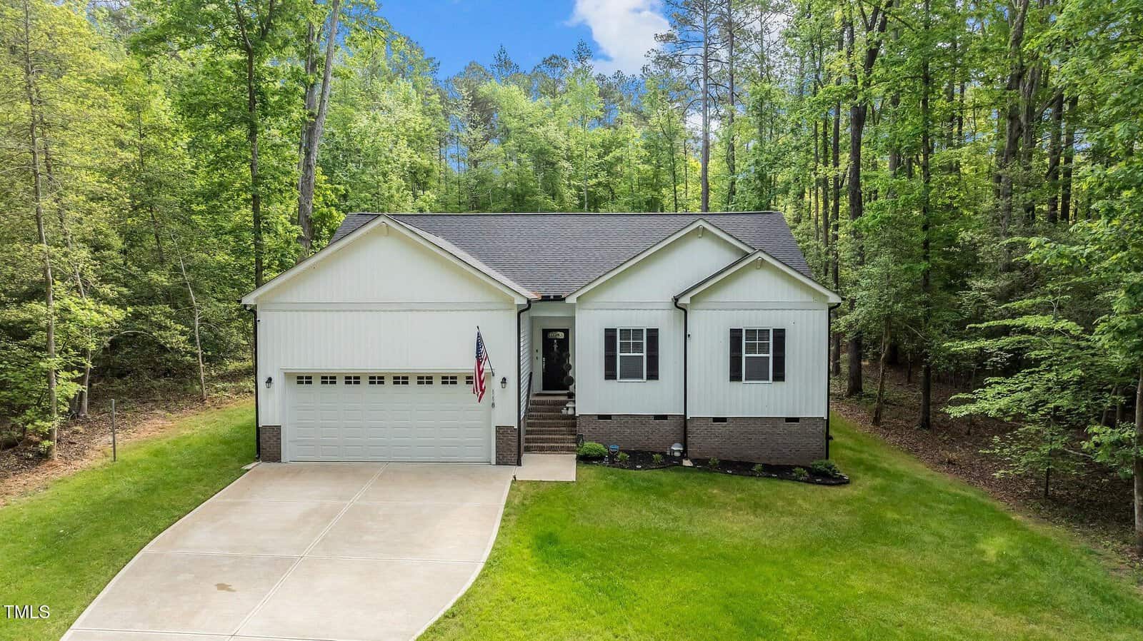 Single-story white house with a gray roof, double garage, American flag, and front steps, surrounded by tall green trees and a neatly mowed lawn, viewed from above on a clear day.
