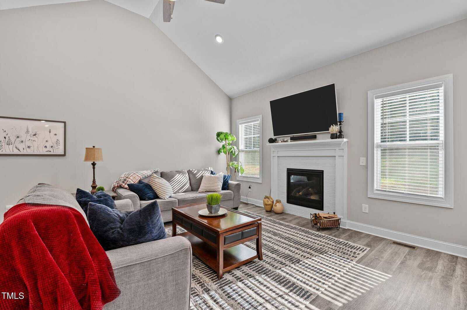 A modern living room with gray sofas, a coffee table, a fireplace with a TV above it, two windows, a potted plant, and a striped rug on wood flooring. Walls are light gray and decor is simple.