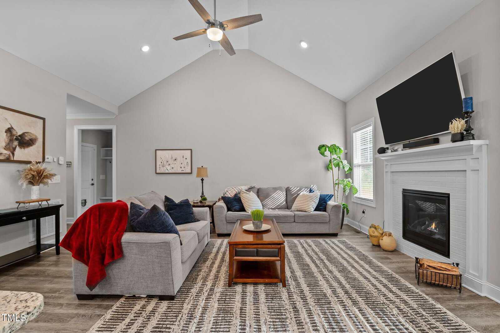 A living room with two gray sofas, a wooden coffee table, a fireplace with a TV above, a patterned rug, houseplants, and neutral decor. The ceiling is vaulted with a ceiling fan and recessed lighting.