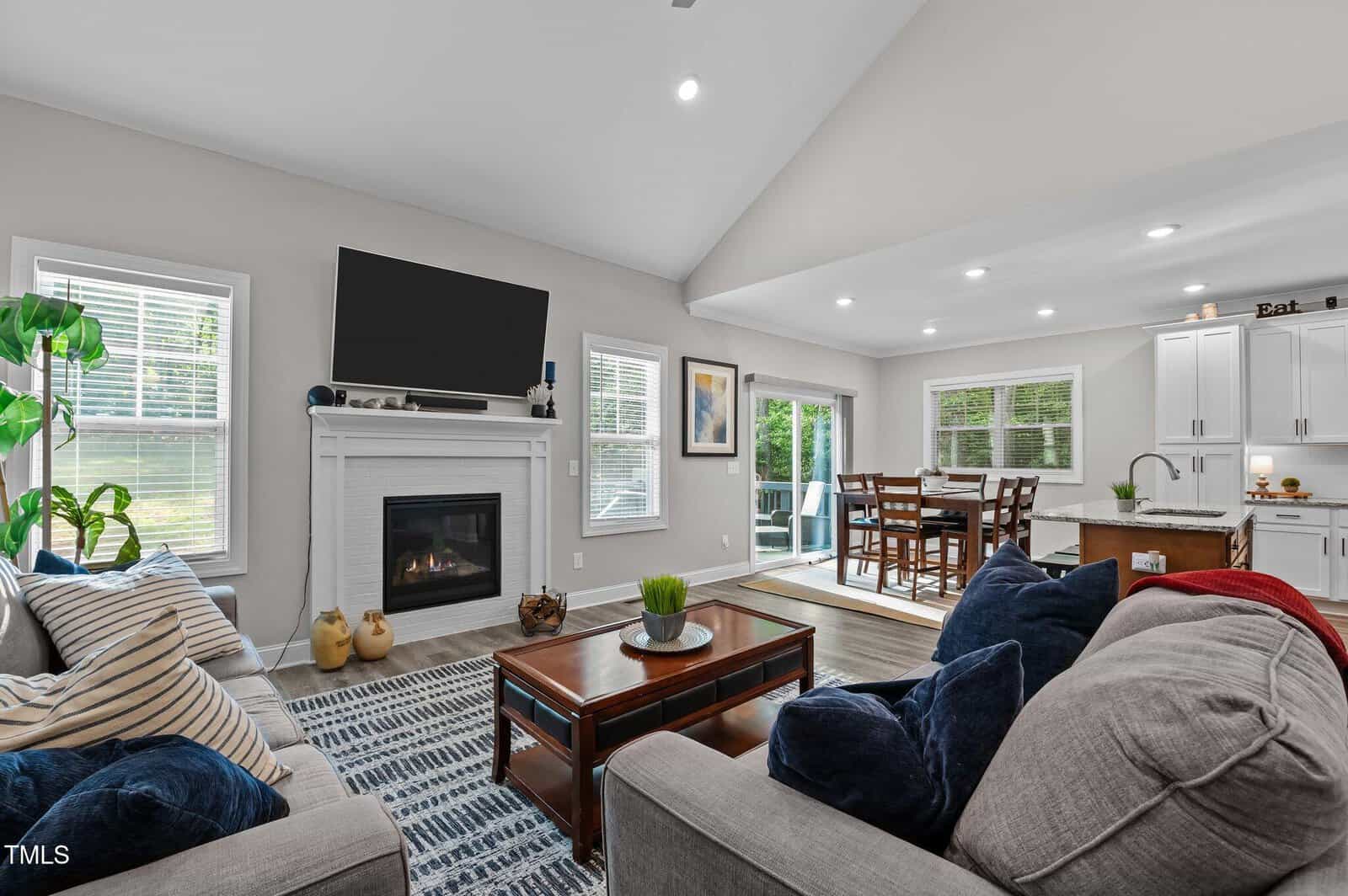 A bright, modern living room with a fireplace, mounted TV, gray sofas with blue and red pillows, a coffee table, and large windows. The open layout leads to a dining area and kitchen with white cabinets.