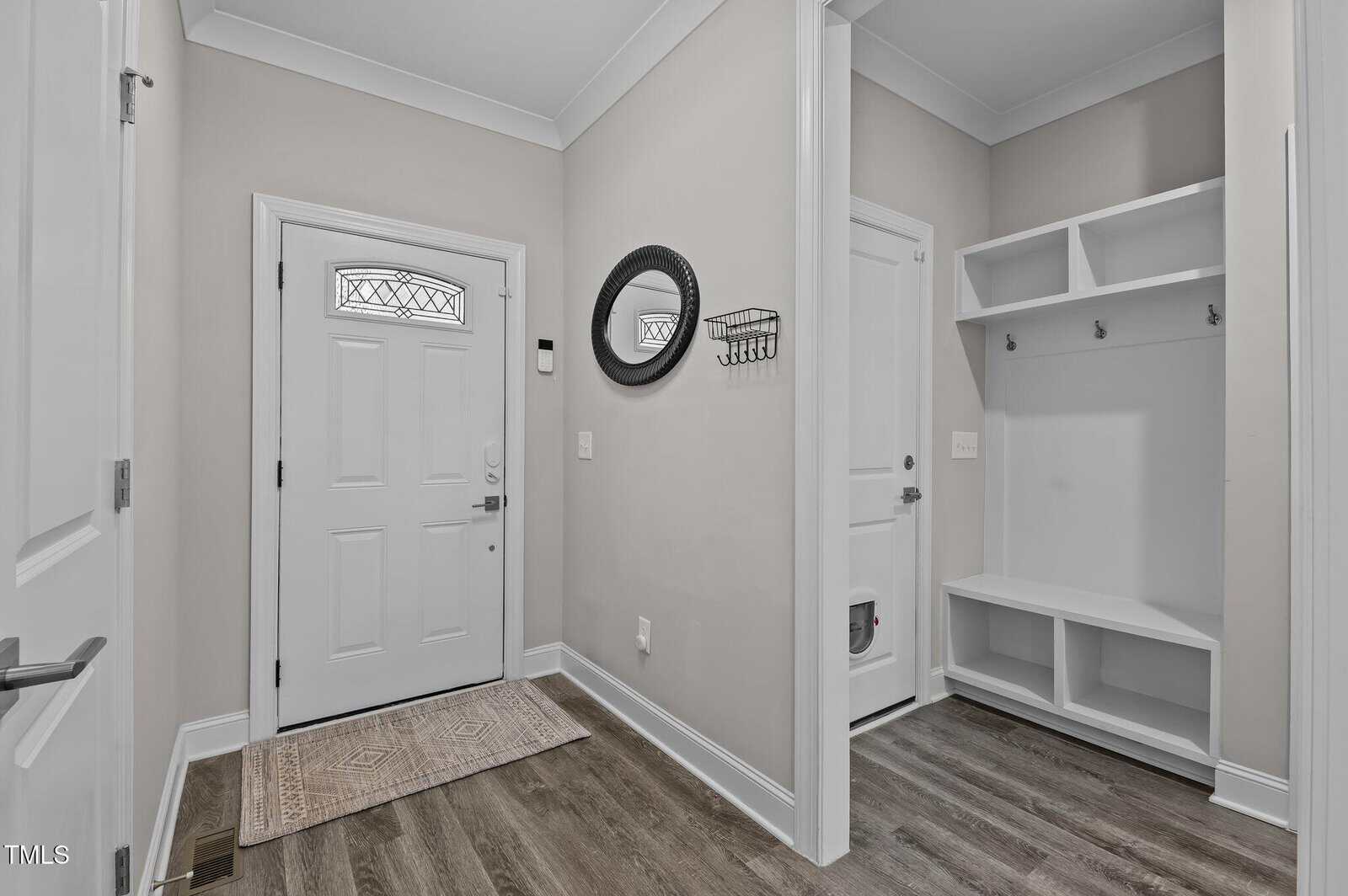 A modern entryway with a white front door, decorative glass window, round mirror, wall hooks, built-in bench with cubbies, and wood-style flooring. Neutral walls and trim create a clean, welcoming look.
