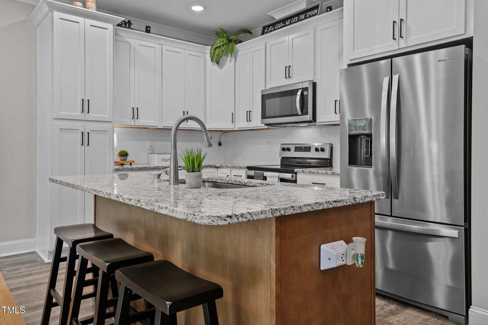 Modern kitchen with white cabinets, stainless steel appliances, granite island with sink and faucet, three black stools, a small potted plant, and wooden floor. An outlet and sanitizer are mounted on the side of the island.