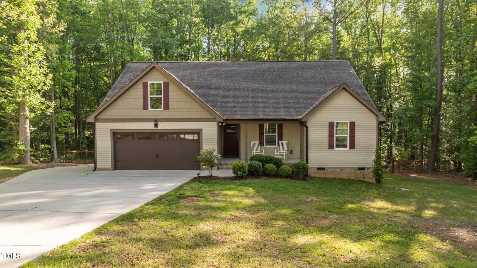 Nestled among tall trees in Kittrell, 4005 Colleen Way features a single-story beige house with brown shutters, a dark garage door, a cozy front porch with two white chairs, and a concrete driveway leading to the garage.