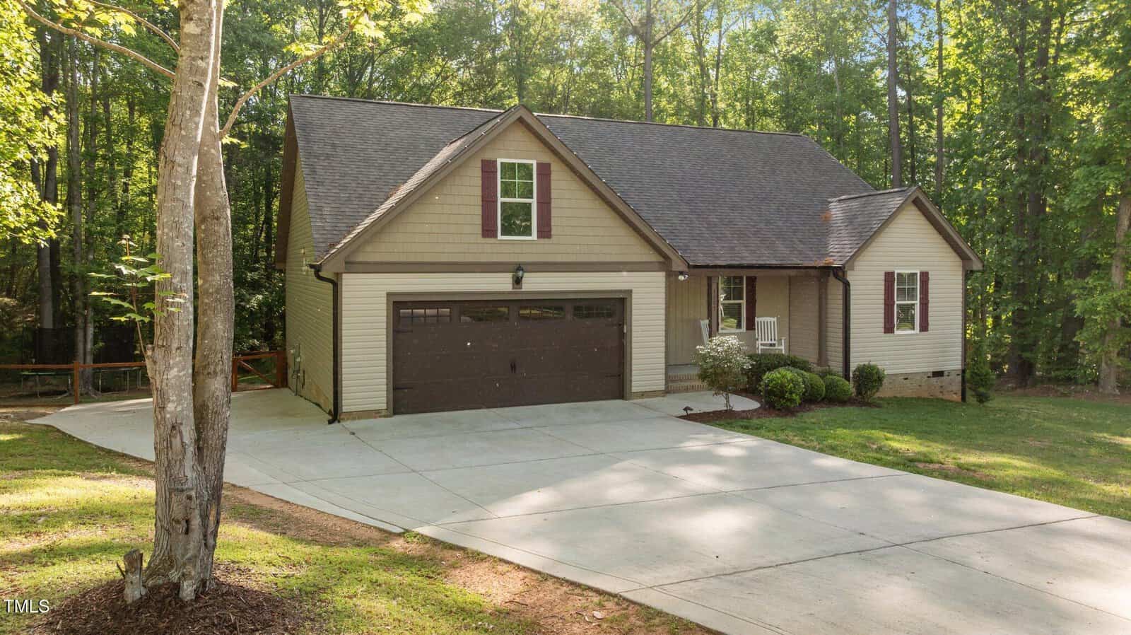 A single-story house with beige siding, dark brown trim, and a two-car garage sits on a large concrete driveway, surrounded by grass and trees on a sunny day.