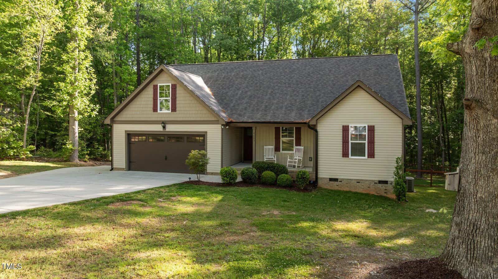 Single-story beige house with dark roof, attached two-car garage, front porch with two white rocking chairs, landscaped yard, and surrounded by tall green trees. Concrete driveway on the left side of the image.