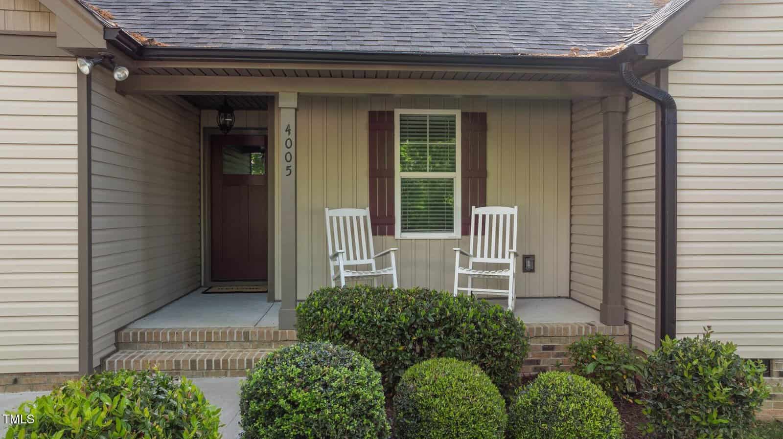 A small porch with two white rocking chairs, a front door to the left, a window with brown shutters, and neatly trimmed bushes in front of the steps. The house number 4005 is displayed next to the door.