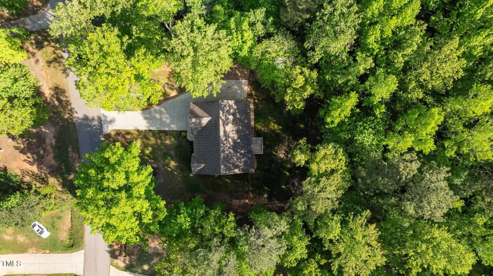 Aerial view of a single house surrounded by dense green trees with a driveway connecting to a residential street; one white vehicle is parked on the street nearby.