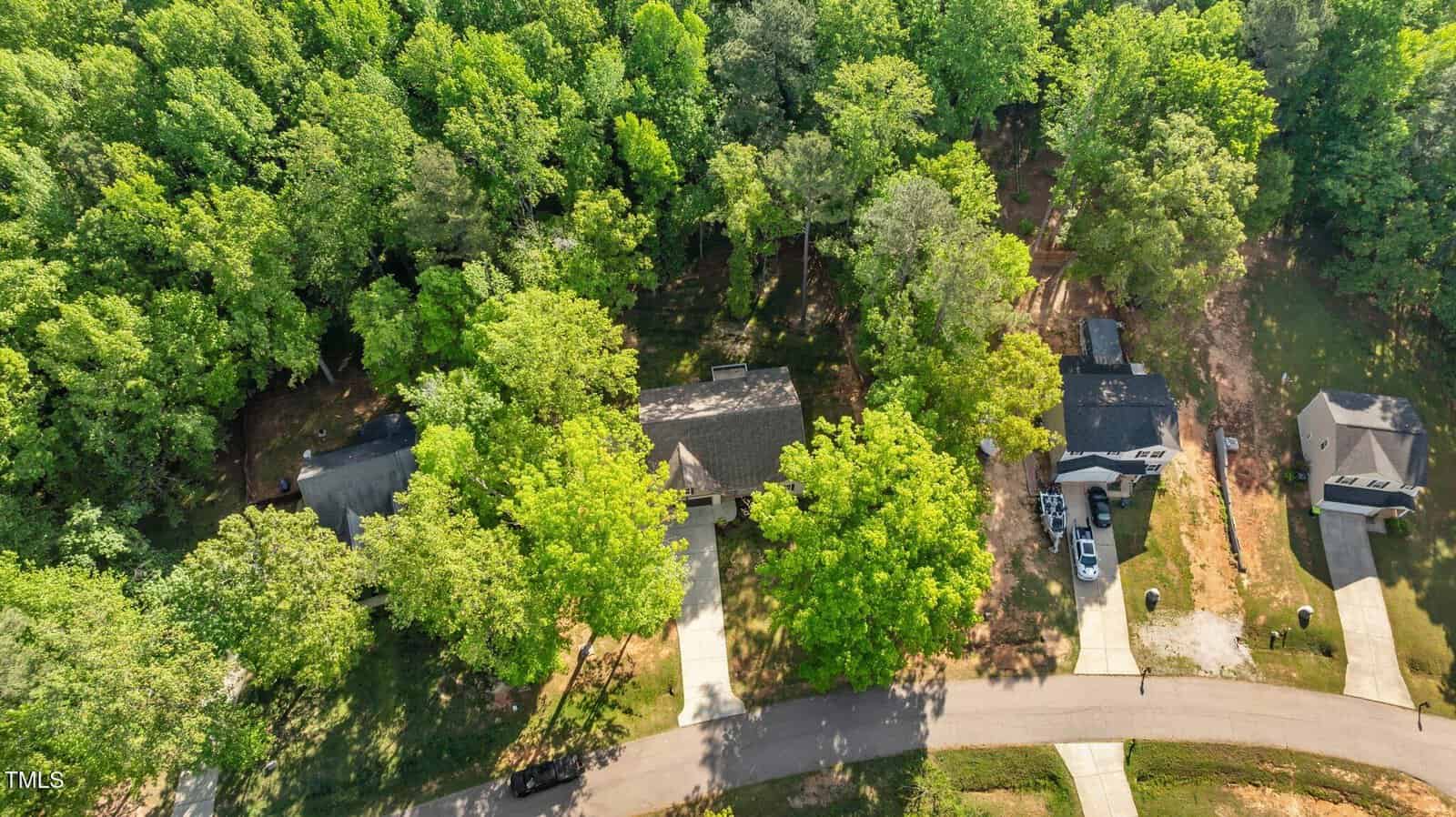 Aerial view of three houses in a suburban neighborhood, each with driveways and surrounded by dense green trees. The street curves in front of the homes, and the area appears quiet and residential.