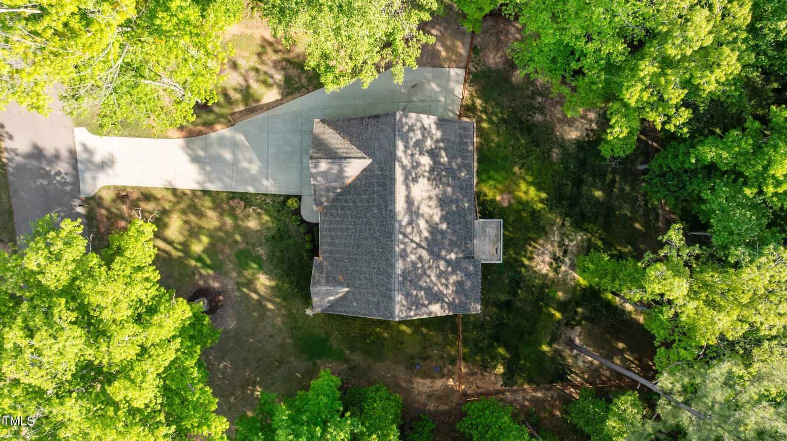 Aerial view of a house surrounded by trees with a curved driveway leading to a road, and a shaded yard with patches of grass and sunlight.