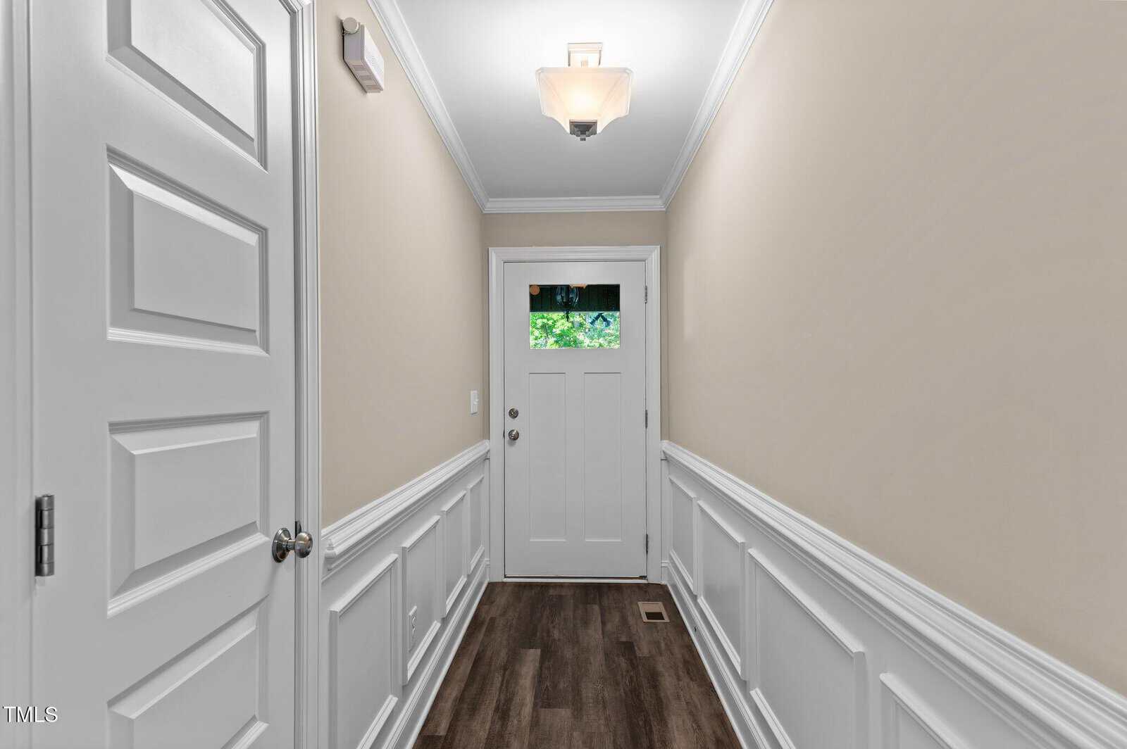 A narrow hallway with beige walls, white wainscoting, dark wood flooring, and a white front door with a glass window at the top. A ceiling light fixture is centered above the hallway.