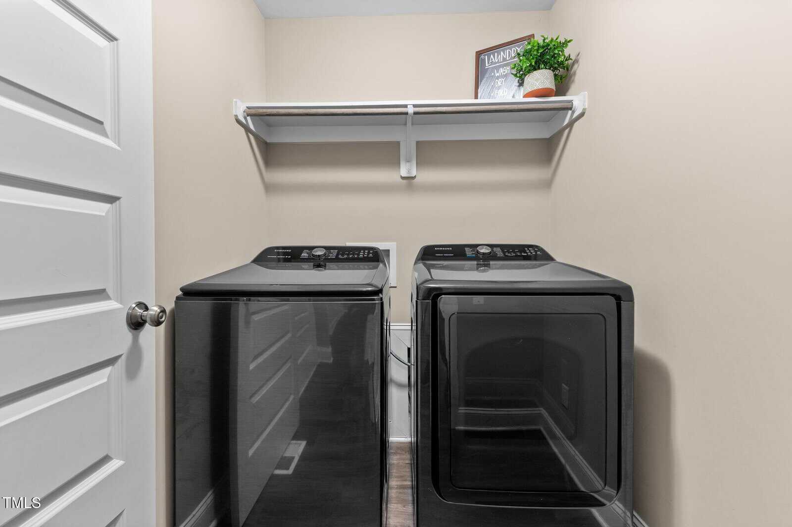 A laundry room with a black washing machine and dryer side by side, a white shelf above holding a framed sign and a small potted plant, and beige walls.