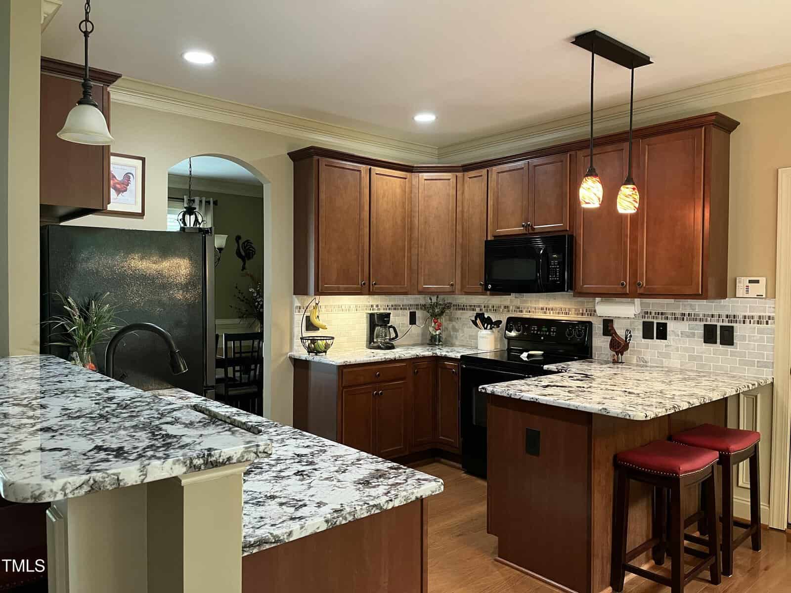 Modern kitchen with brown cabinets, marble countertops, black appliances, pendant lights, and two red barstools. A dining area with a table and plant is visible through the arched doorway.