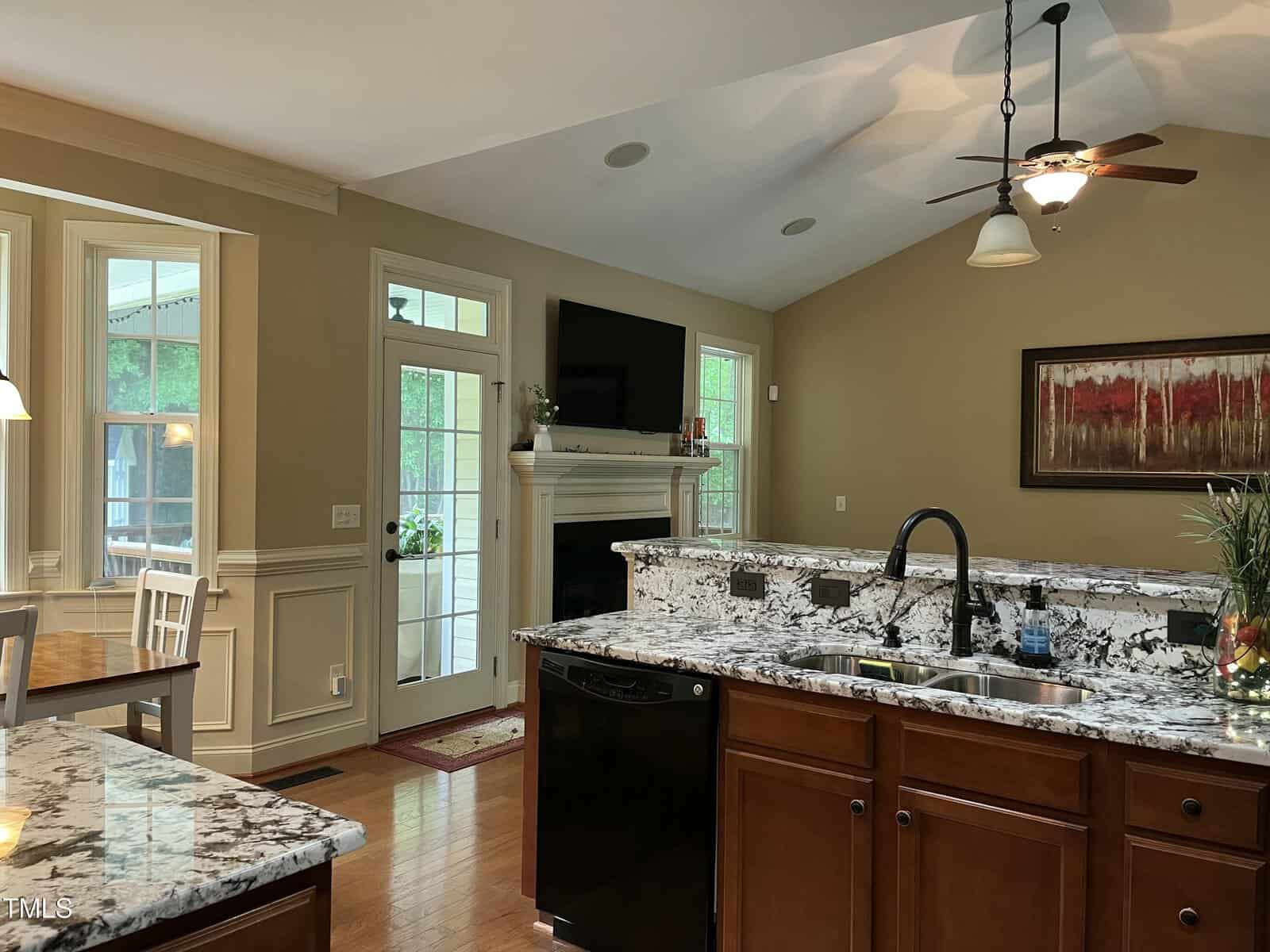 Open kitchen with granite countertops and dark appliances overlooks a living area with a wall-mounted TV, fireplace, and large windows. Dining nook with table and chairs is visible to the left. Ceiling fans and recessed lighting are present.