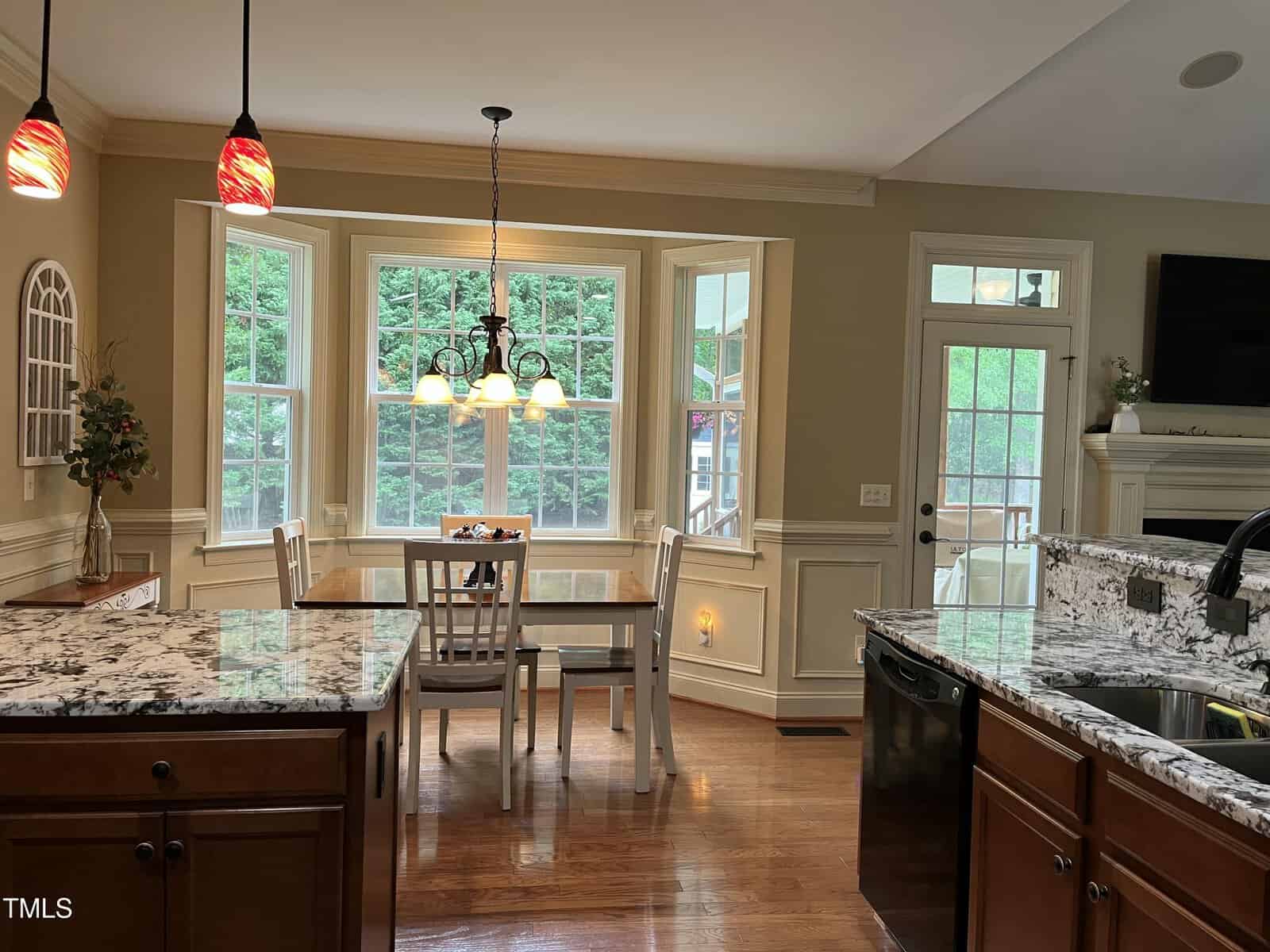 A kitchen with granite countertops, wooden cabinets, a sink, and a dishwasher. A dining table with four chairs sits in a bay window area. Pendant lights hang above the counter; greenery is visible outside the windows.
