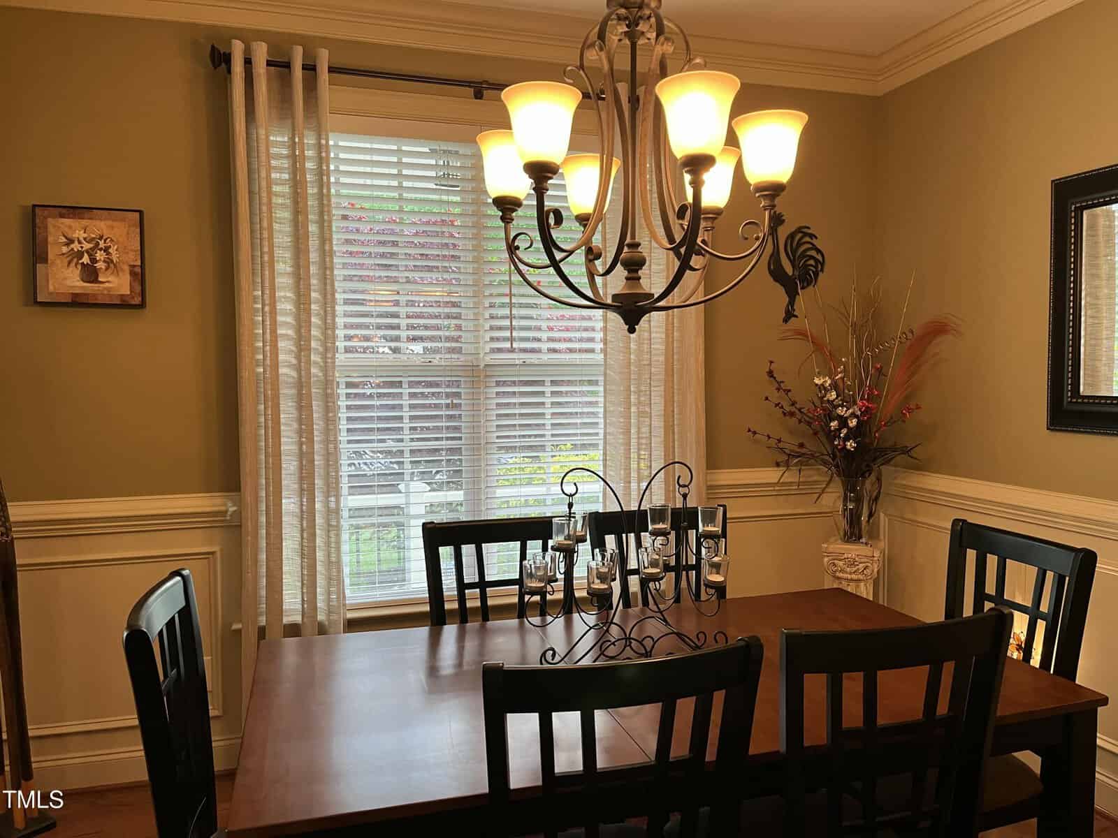 A dining room with a wooden table, six black chairs, a decorative black candle holder, a chandelier with lit bulbs, a window with white curtains, a framed picture on the wall, and a tall vase with artificial flowers.