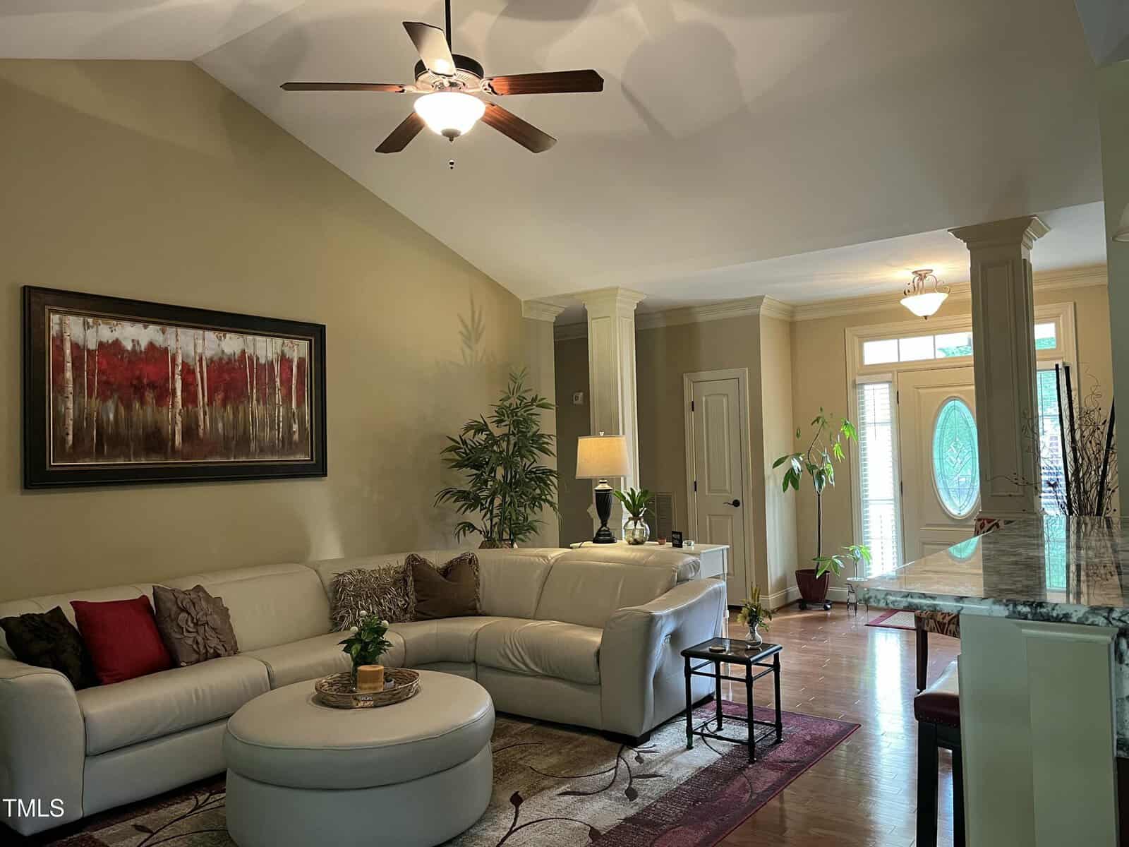 A living room with beige walls and wood flooring features a white sectional sofa, round ottoman, and red-accented decor. A ceiling fan and modern artwork are present, and large windows by the entrance let in natural light.