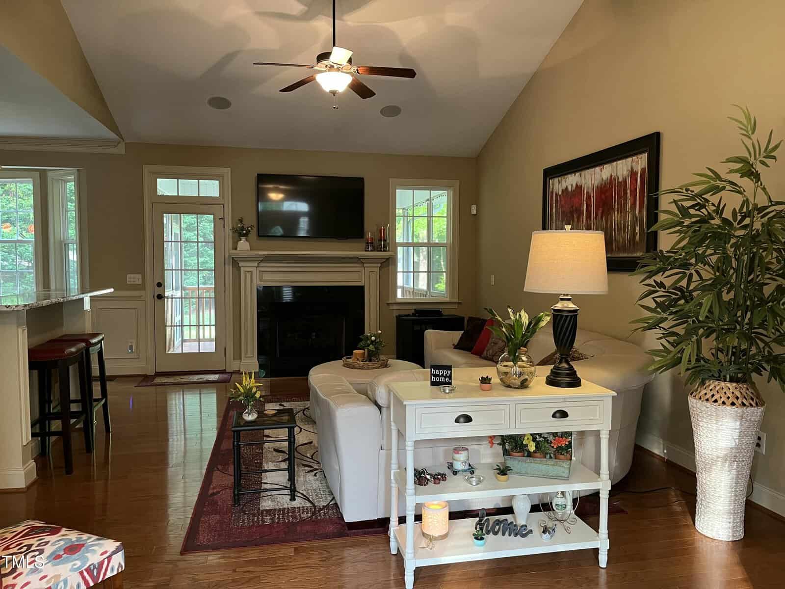 A living room with wood floors, beige walls, a white couch, a side table with a lamp, a TV above a fireplace, a plant, bar stools at a counter, and large windows and doors letting in natural light.